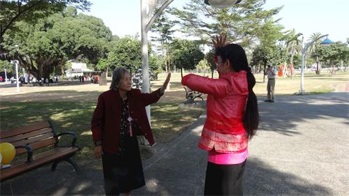 '圖8：現年八十幾歲的蔡老太太（左），巧遇法輪功集體活動，她走近了煉功隊伍，展開雙臂跟著學煉，旁邊學員熱心的教她煉功。'